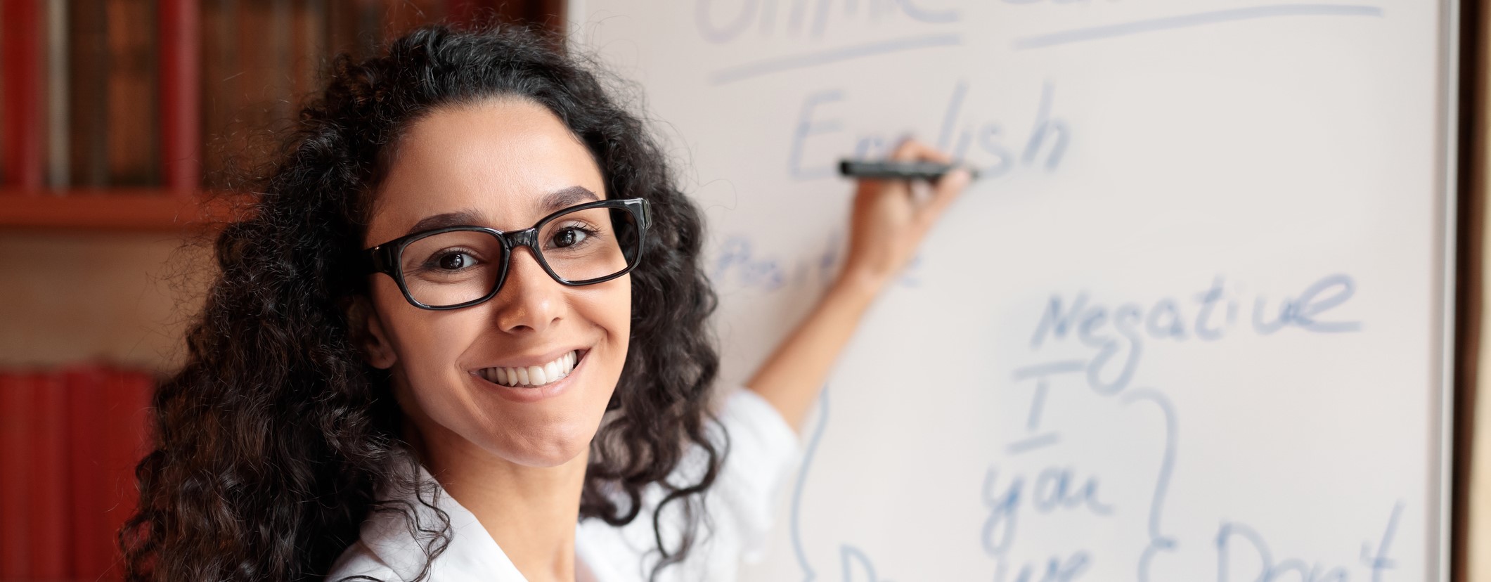 Portrait of smiling female teacher standing at whiteboard, explaining grammar rules to students.